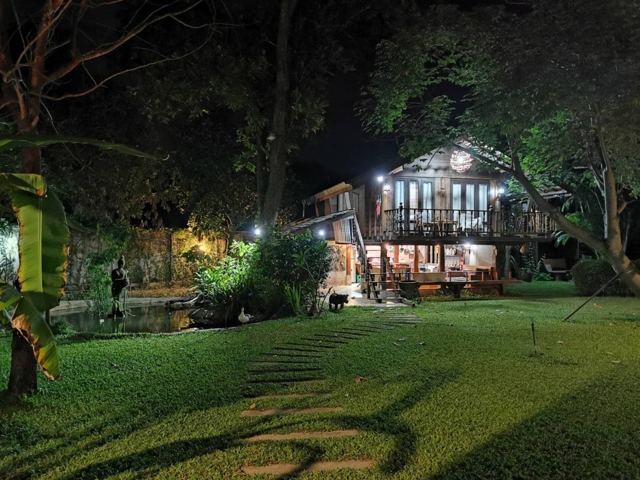 บ้านไม้ใต้ถุนสูงท่ามกลางสวนยามค่ำคืน แสงไฟอบอุ่นและบรรยากาศเงียบสงบ