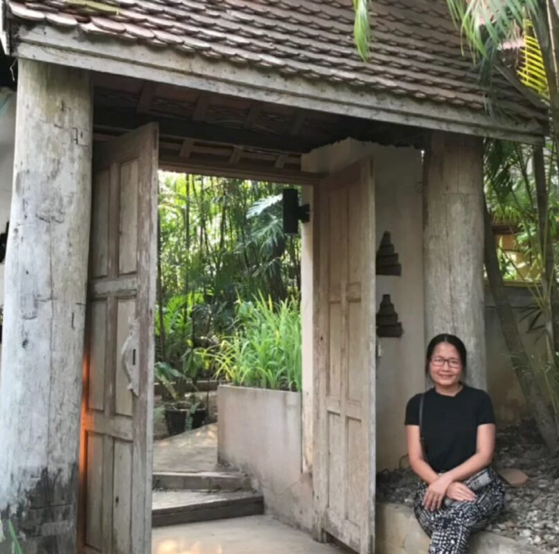 ภาพถ่ายจริงของเจ้าของที่พักนั่งอยู่บริเวณประตูไม้ท่ามกลางสวน
